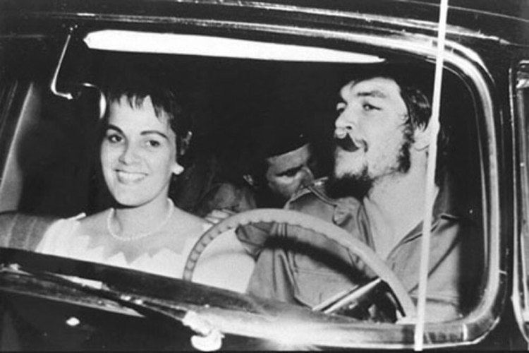 Aleida March smiling while Che Guevarra smoking with a man inside a car. Aleida wearing a pearl necklace and a white dress while Che with a beard and wearing a long sleeve.