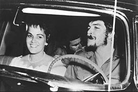 Aleida March smiling while Che Guevarra smoking with a man inside a car. Aleida wearing a pearl necklace and a white dress while Che with a beard and wearing a long sleeve.