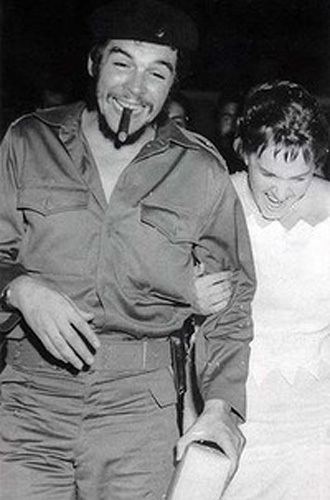 Aleida March and Che Guevarra are laughing together while Che is smoking. Aleida wearing a white gown while Che wearing a black hat, long sleeves, and pants.
