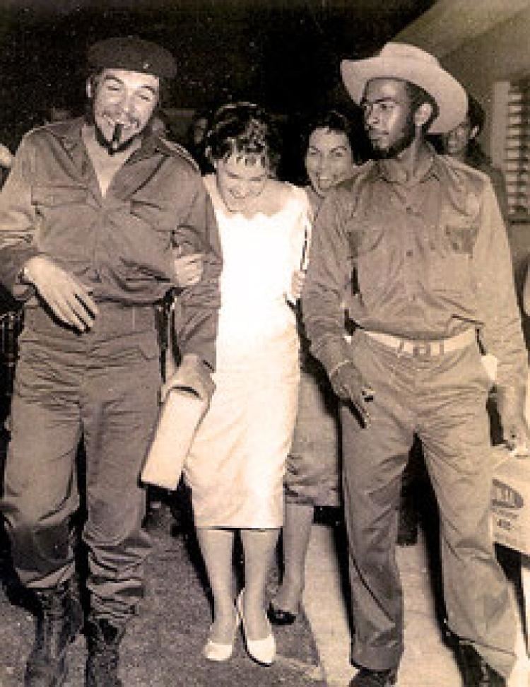 Aleida March and Che Guevarra are laughing together while Che is smoking and surrounded by their guests. Aleida wearing a white gown while Che wearing a black hat, long sleeves, and pants.