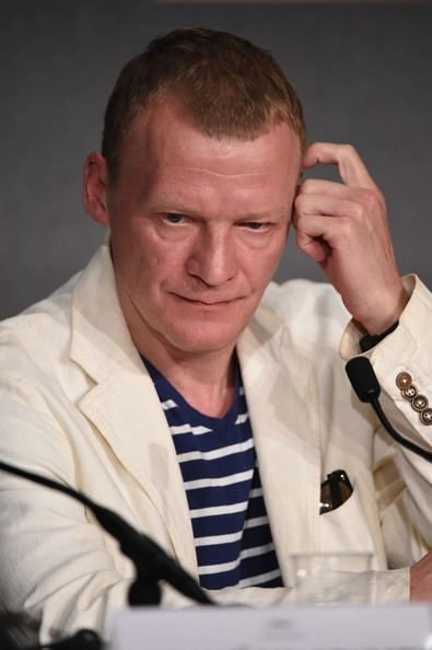 Aleksei Serebryakov scratching his head while wearing cream coat and striped shirt