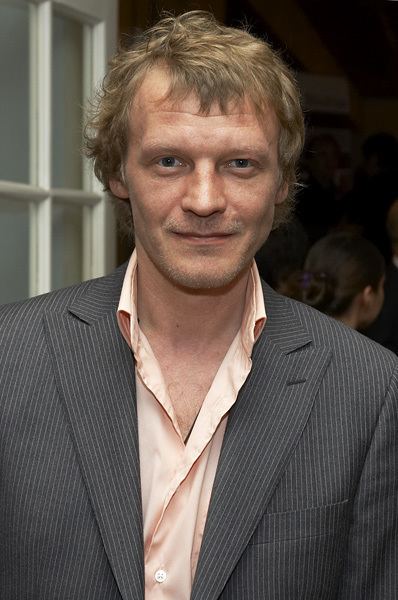 Aleksei Serebryakov smiling and wearing striped coat and orange long sleeves