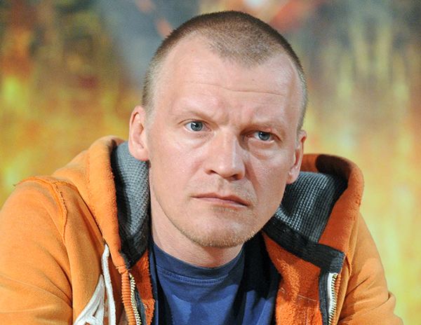 Aleksei Serebryakov looking afar while wearing orange jacket and blue shirt