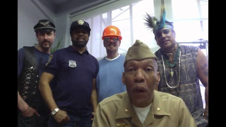Alex Briley at the front speaking and wearing beige long sleeves and a beige hat together with four other members of the band Village People
