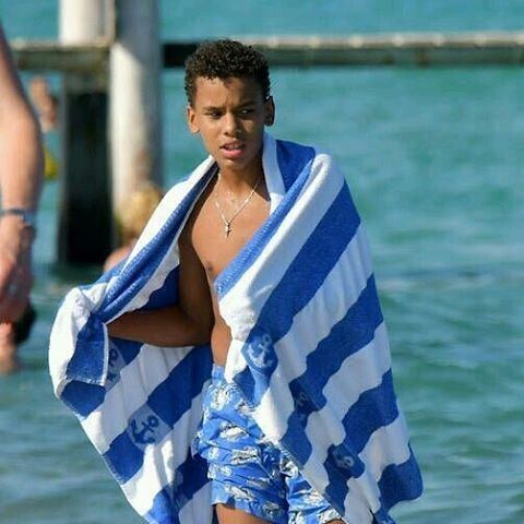 Alexandre Coste wearing blue shorts, a necklace and holding a white and blue towel at the beach.