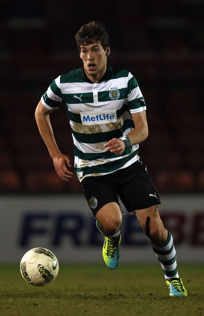 Alexandre Guedes playing football with curly hair and wearing a stained shirt with green and white stripes, black shorts, knee socks with green and white stripes, and blue and yellow-colored shoes