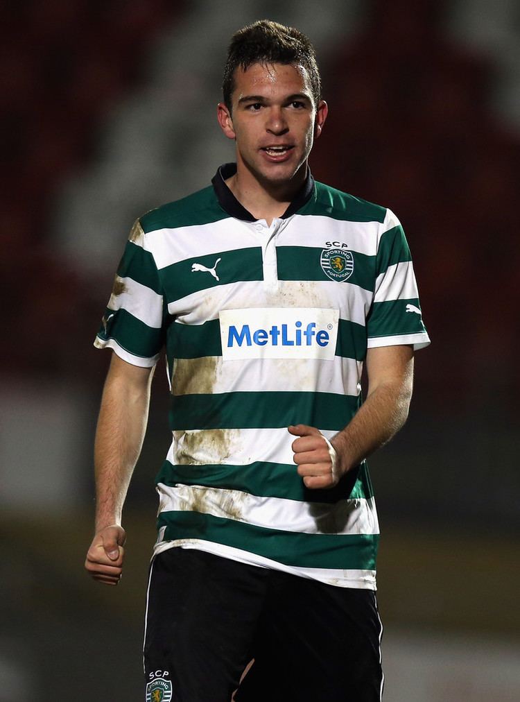 Alexandre Guedes looking happy and clenching his fists during a football game while wearing a stained shirt with green and white stripes and logos written on it and  black shorts
