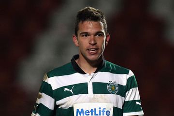 Alexandre Guedes looking happy  during a football game while wearing a stained shirt with green and white stripes and logos written on it