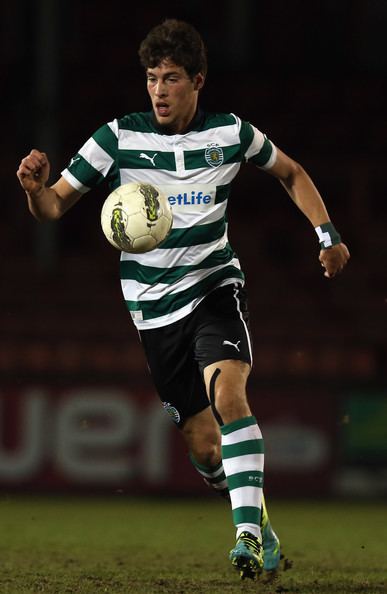 Alexandre Guedes running and acting to kick the ball on air during a football game and wearing a shirt with green and white stripes, black shorts, knee socks with green and white stripes, and blue and yellow-colored shoes