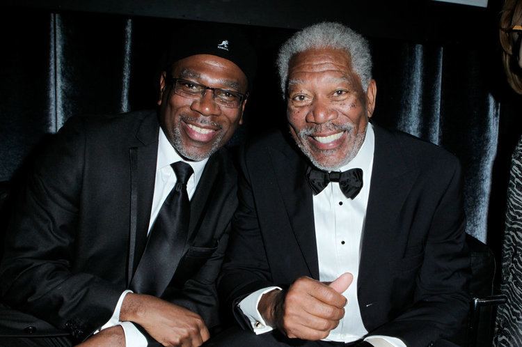 Alfonso Freeman smiling with his father Morgan Freeman while wearing a black coat, white long sleeves, black necktie, and eyeglasses