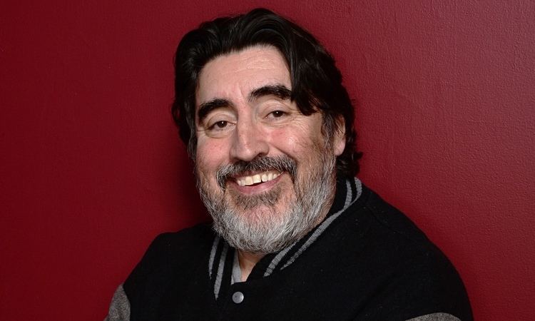 Alfred Molina smiling with a mustache and beard while wearing a gray and black jacket