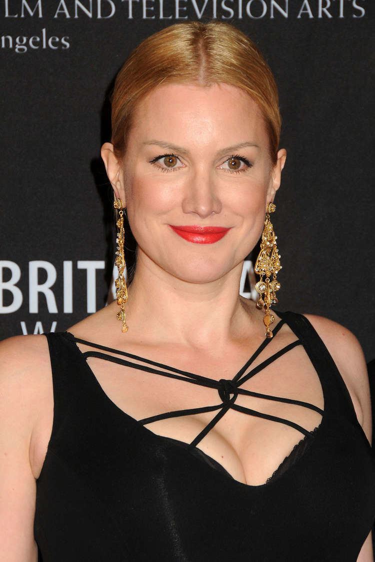 Alice Evans smiling, is an American-English actress during the 2011 Britannia Awards. She has brown eyes, and red lips, wearing a pair of gold and diamond dangle earrings, in a black sleeveless and crisscrossed-straps gown exposing her cleavage.