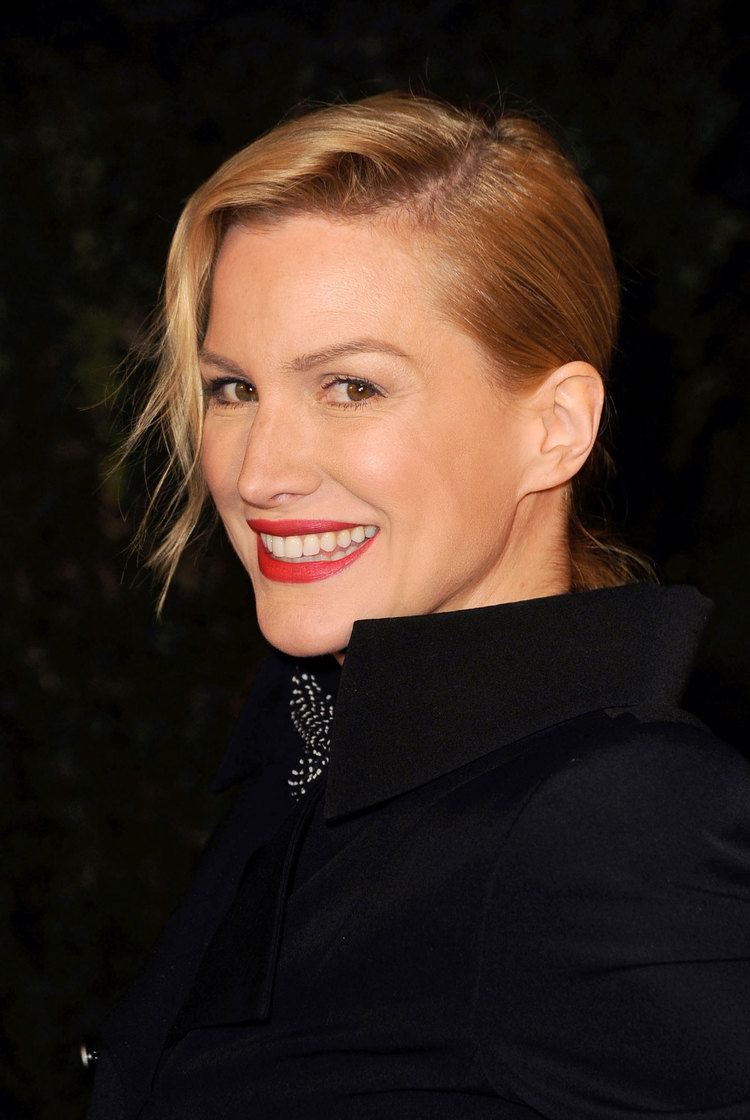 Alice Evans smiling in sideways, an English-American actress with brown eyes, red lips, blonde hair tied with side bangs, wearing a polka dot neck scarf, black inner blazer, and a midnight-blue coat.