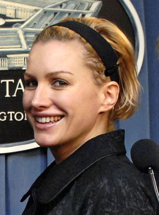 Alice Evans smiling and looking sideways, an American-English actress during a tour of the Pentagon in 2006. She has short blonde hair, a black headband, and a black hair clip with a microphone in front, wearing a black laced-collared top.