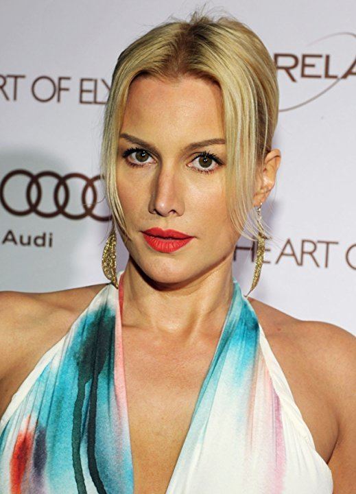 Alice Evans looking fierce is an American-English actress during the 2012 Art of Elysium Heaven Gala with red lips, her blonde hair tied up and bangs on the sides, wearing gold dangling earrings, and a deep-V watercolor print halter gown.