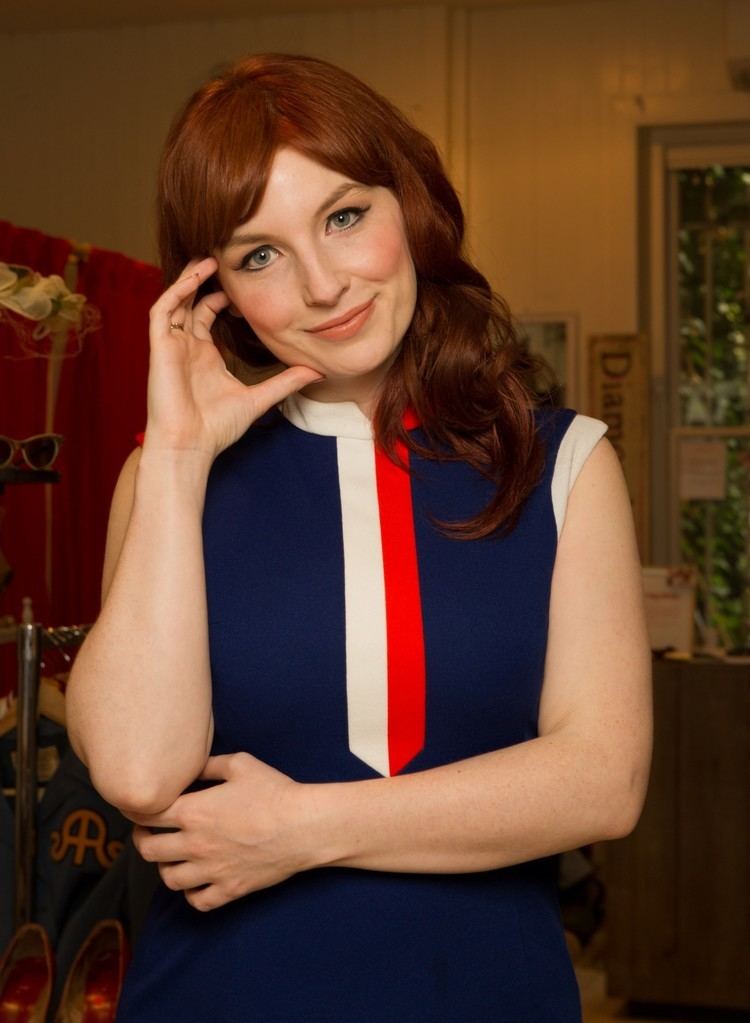 Alice Levine, on her hands-on-face pose, wearing a sleeveless dress inside a room