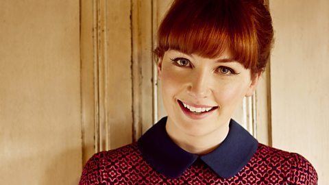 Alice Levine smiles while wearing a red collared shirt