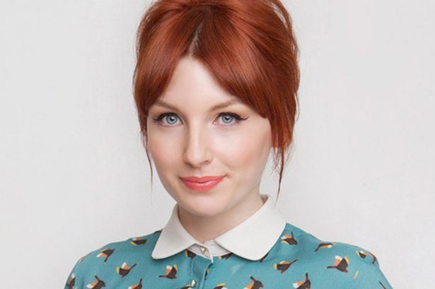 Alice Levine, in her reddish hair, smiles while wearing a blue collared shirt