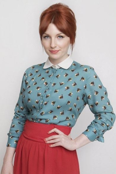 Alice Levine smiles while wearing a blue collared long-sleeved shirt and a red skirt