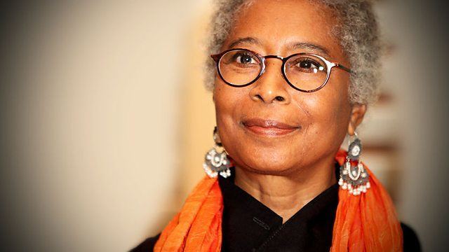 Alice Walker is smiling, standing in a white room, has white-gray hair wearing black eyeglasses a gray-white dangling earrings, an orange scarf, and black top.
