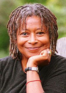 Alice Walker is smiling, sitting leaning with her left elbow on a wooden bench outside with greenery at the back, she has gray-brow thick hair wearing a black long sleeve, silver dangling earrings, has silver ring and black with silver watch and a red strap on her left arm.