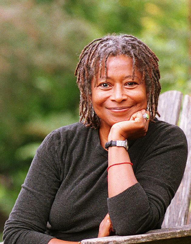 Alice Walker is smiling, sitting leaning with her left elbow on a wooden bench outside with greenery at the back, she has gray-brow thick hair wearing a black long sleeve, silver dangling earrings, has silver ring and black with silver watch and a red strap on her left arm.