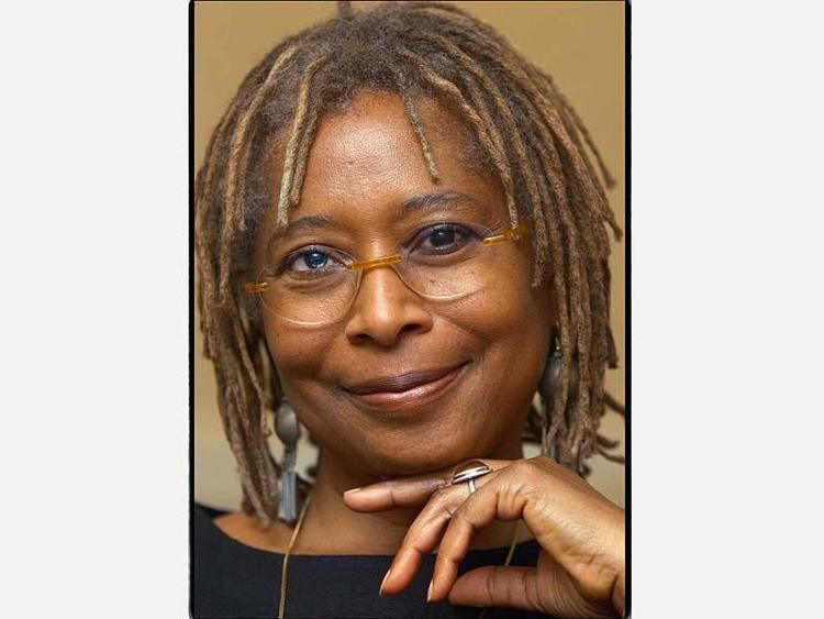 Alice Walker is smiling, has brown thick hair wearing eyeglasses, a gray long earrings, a gold neck lace and a large silver ring. Behind is a brown wall.