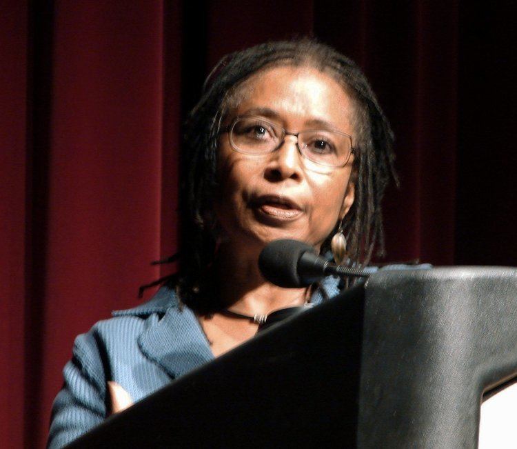 In a conference room with a large red curtain at the back, Alice Walker is serious, speaking through the mic on a black host table, has black thick hair wearing eyeglasses, long earrings, a black necklace and a blue corduroy coat.