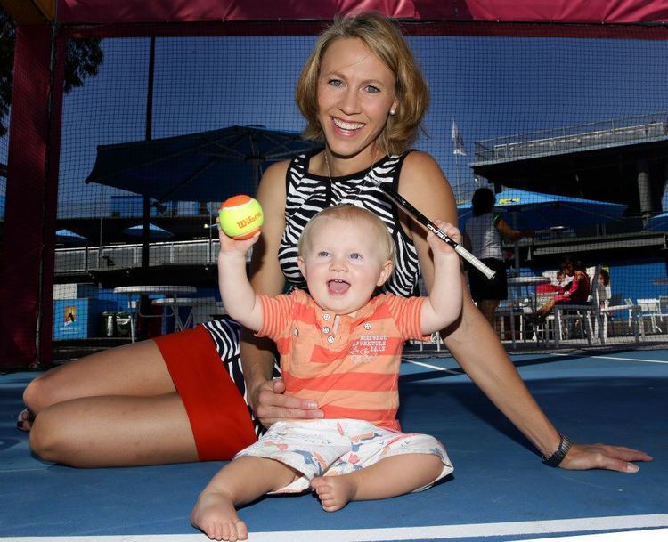Alicia Molik smiling with the baby in front of her while she is wearing a black and white top and red skirt
