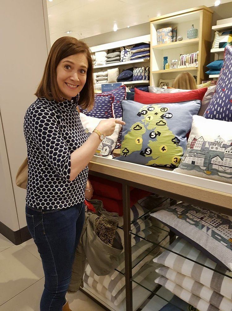 Alina Jenkins smiling while pointing at the pillow, with short blonde hair, wearing a black and white polka dots blouse, and blue pants.