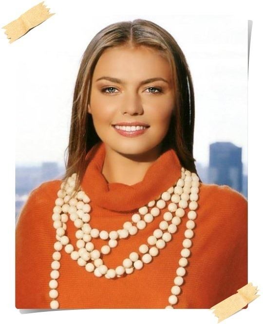 Alina Kabaeva smiling, with straight blonde hair, wearing a pearl necklace and an orange turtleneck top.