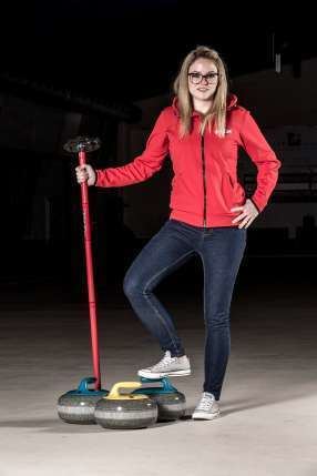 Alina Pätz smiling while holding a curling broom with curling stones on the floor, with blonde hair, wearing eyeglasses, a red jacket, black pants, and white sneakers.
