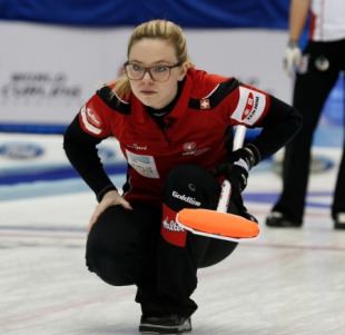 Alina Pätz doing curling with a serious face and holding a curling broom, with blonde hair, wearing eyeglasses, a multi-colored jacket, black pants, and black shoes.