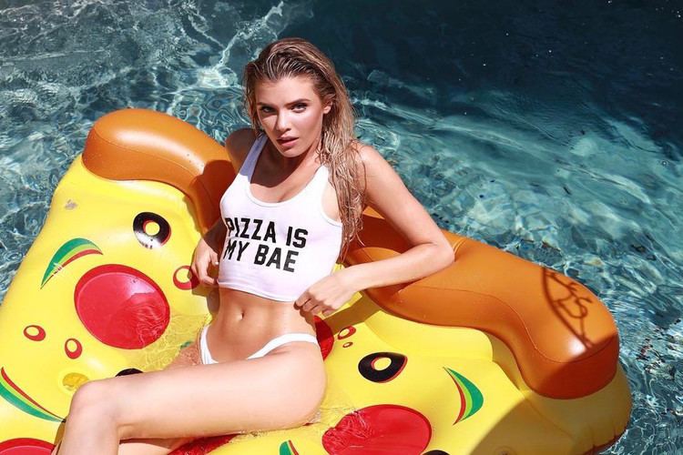 Alissa Violet with a fierce look while lying on a pizza-style inflatable floater, with wet blonde hair, and wearing a white two-piece swimsuit.