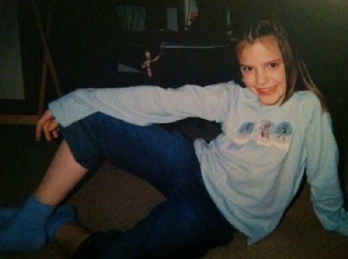Young Alissa Violet smiling while sitting on the floor, with her hands on her leg, wearing a light blue sweatshirt, blue jeans, and blue socks.