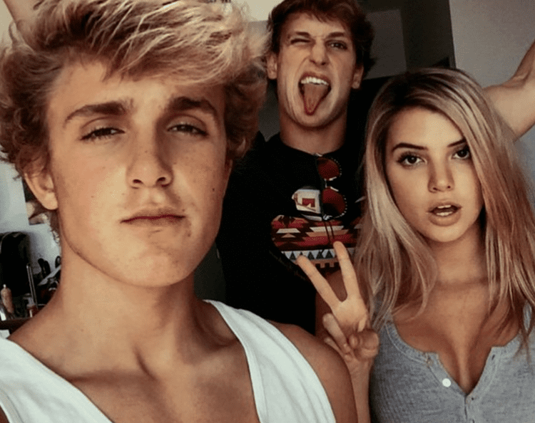 Alissa Violet with Jake and Logan Paul while doing a peace sign. Alissa with blonde hair and wearing a gray top, Jake wearing a white sleeveless shirt, and Logan spitting his tongue out and wearing a black shirt.