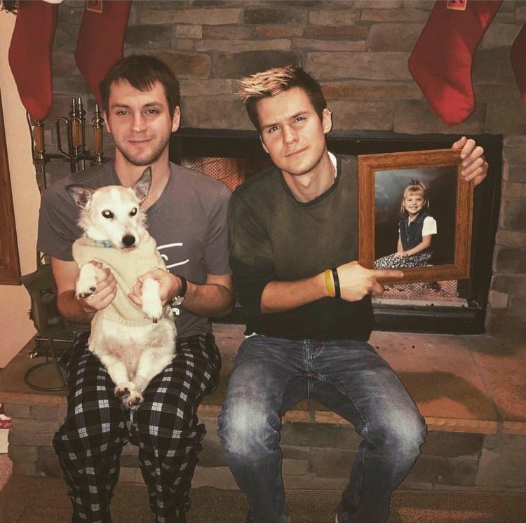 Joshua Butler and Rhett Butler are smiling. Joshua holding a white dog, wearing a gray shirt and a checkered black and white pajama while Rhett holding a picture frame of Alissa, wearing a long sleeve shirt and jeans.