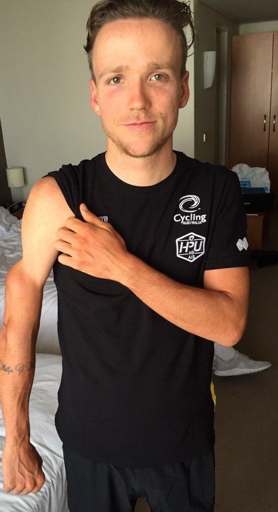 Alistair Donohoe with a tight-lipped smile while showing his arms and wearing a black t-shirt and black shorts