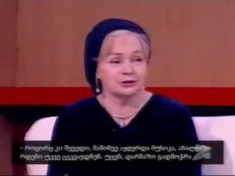 Alla Dudaeva is talking while sitting on a white couch during an interview on the Georgian TV show "Profile". Alla Dudaeva with blonde hair, wearing a dark blue headscarf, earrings, a necklace, and a dark blue dress.
