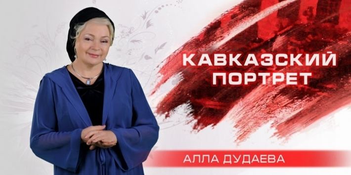On the left, Alla Dudaeva appears on “First Caucasus TV", a Russian-language TV channel. Alla Dudaeva with a tight-lipped smile while holding her hands together, with blonde hair, wearing a black headscarf, earrings, a necklace, and a blue robe over a black shirt.On the right are the Russian words "Caucasus Portrait" and "Alla Dudaeva".