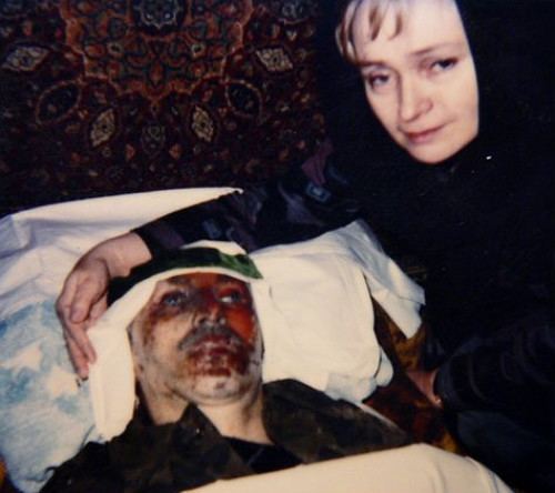 Alla Dudayeva with a sad face while standing beside her husband's dead body with blood over his face. Alla Dudayeva with blonde hair, wearing a black headscarf and a black hijab.