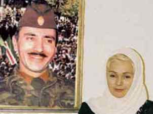 Alla Dudayeva with a sad face standing in front of a picture frame of her husband Dzhokhar Dudayev. Alla Dudayeva with blonde hair, wearing a white headscarf and a black hijab.