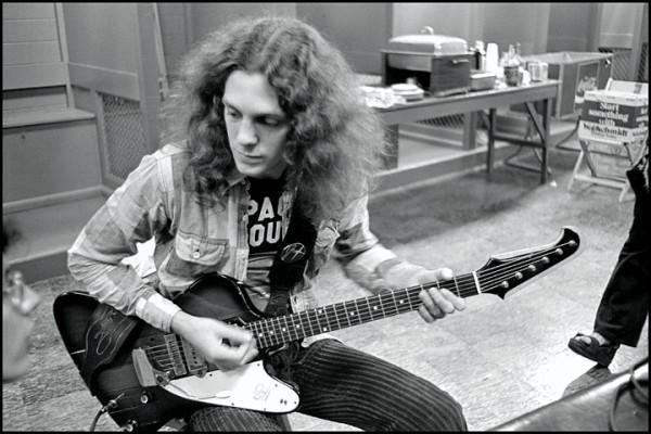 Allen Collins sitting down and playing a guitar on set while wearing a black shirt under a jacket.