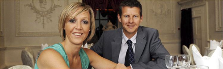 Allison Curbishley and Steve Cram are smiling. Allison with short blonde hair with pearl earrings and wearing a light green sleeveless top while Steve is wearing a gray coat over white long sleeves and a black necktie.