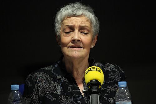 Amaya Uranga is serious, has white hair, mouth half opened, both eyes closed, in front of a yellow microphone, with two water bottles on both sides, wearing silver earrings, and a black top with a white design.