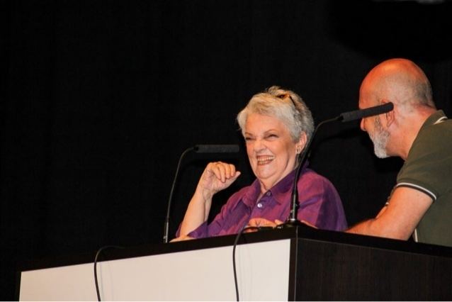 A woman (left) is smiling, has white hair, her right hand up on the white-brown table, and a microphone in front, wearing a ring on her right hand, glasses on her head, earrings, and a violet top. A man (right) is bald, has a black and white mustache, his hands on the white-brown table, and a microphone in front, wearing a green top with white stripes on the shoulder.