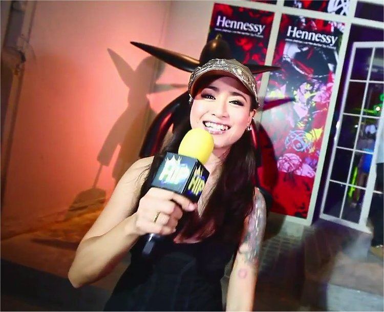 Ammara Siripong smiling while holding a microphone and wearing a black tank top and gold cap