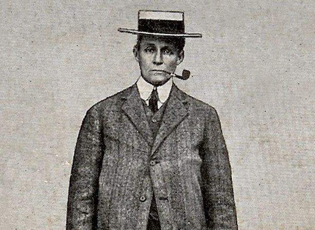 Amy Bock is serious, standing while using a pipe cigarette, wearing a black and white hat, white long sleeves, and a black necktie under a black suit with buttons.
