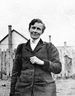 Amy Bock is smiling and has black short hair at the back of a house in the background in an old photo, she is wearing a white polo under a black coat