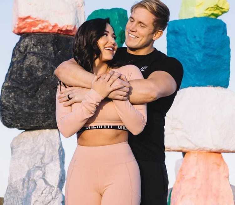 Ana Cheri smiling in a pink sports outfit while being embraced by his husband Ben Moreland wearing a black shirt